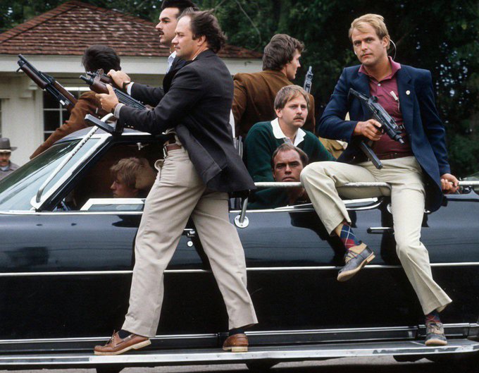 A vintage film still showing several men in formal attire holding firearms while sitting in or standing on a black convertible car.