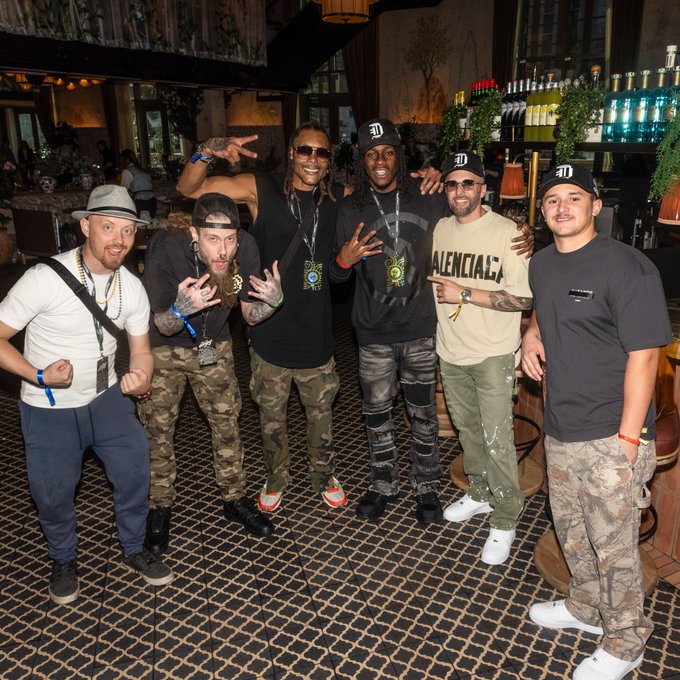 Six men pose together in a restaurant/bar setting, including one wearing a 'Balenciaga' shirt and several wearing caps with a 'D' logo, with lanyards around their necks.