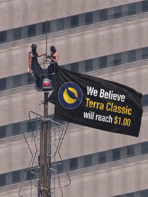 Two people hanging a banner from a tall pole that reads 'We Believe Terra Classic will reach $1.00' with the Terra logo.