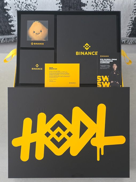 A Binance-branded black gift box with the word 'HODL' spray-painted in yellow on the front, containing merchandise items and a yellow playing card.