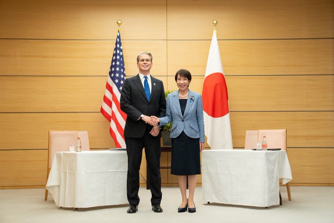 Two officials—one in a U.S. flag suit, the other standing before the Japanese flag—shake hands in a formal setting, symbolizing U.S.-Japan diplomatic discussions ahead of Japan's anticipated September 18 interest rate decision.