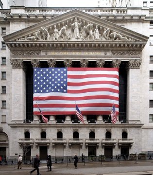 The neoclassical facade of the New York Stock Exchange building is shown, featuring Corinthian columns and a pediment sculpture, with a large American flag draped across the front entrance.