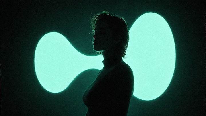 A moody silhouette photo of a person standing in profile against a glowing teal, bean-shaped light installation in a dark setting.
