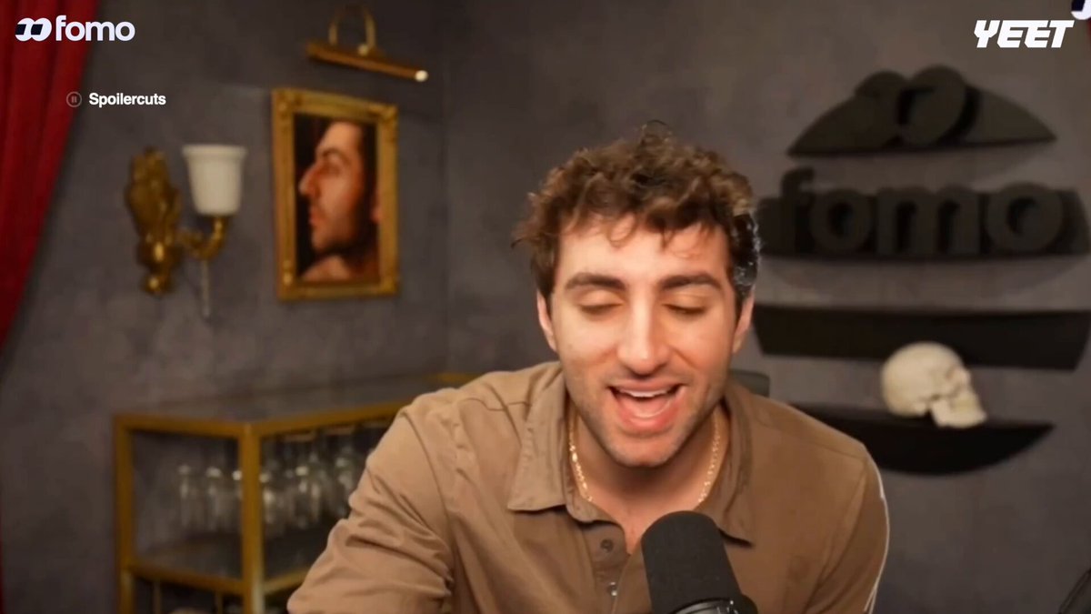 The image features a man sitting at a microphone, smiling as he speaks into it. The background of the image shows a gray wall with a painting hanging above him. There are also two wine glasses placed near the edge of the frame on the right side of the photo. The caption for this image reads "Hyperliquid is the SpaceX of Finance." This phrase suggests that Hyper Liquid, a company named after the founder of SpaceX, is making waves in the finance industry with its innovative approach to investing and raising capital.