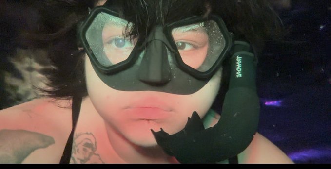 A close-up of a person wearing a snorkel mask with "UNIQUE" printed on the snorkel, their face partially visible with dark tousled hair and a tattoo peeking from a tank top strap. The mask's tinted lenses and underwater aesthetic visually play on the "Buy the dip" message, evoking the imagery of diving deep into a market downturn to scoop up deals.
