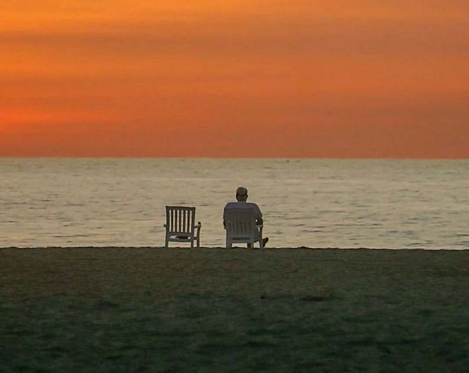 A serene silhouette of a person sitting in a chair facing the ocean at sunset, with an empty chair beside them on a grassy shore.