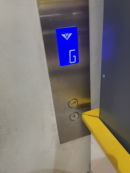 The image shows a gray metal elevator door with a digital display that reads "G". The door has two buttons, one of which is yellow. There are no people visible inside the elevator at the moment.