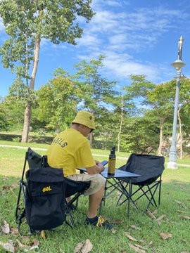 A man wearing a yellow shirt and cap sits in a camping chair in a park using a laptop, with a second empty chair next to him, surrounded by trees on a sunny day.