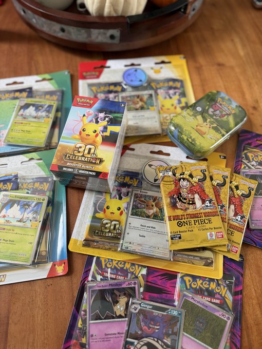 An assortment of Pokémon merchandise on a wooden table, including booster packs, individual cards featuring Eevee and Gengar, a '30th Celebration' booster bundle box, and a metal tin.