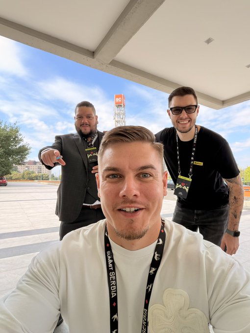 Three men pose for a selfie under a canopy at what appears to be a gaming or crypto event, with the front person wearing a lanyard reading "SQUAD SERBIA" and "NEMESIS" while the man on the right wears a "100x" badge — fittingly complementing the "100x LONG" caption by visually representing bullish optimism through the "100x" branding.