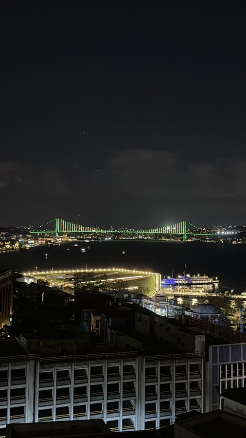 A nighttime aerial view of a cityscape featuring a brightly illuminated green suspension bridge spanning a waterway, with a lit pier and docked ferry below and modern building rooftops in the foreground. The image pairs with a confident post urging followers to act on cryptocurrency advice, suggesting that the nighttime cityscape symbolizes the quiet before a financial breakthrough or hidden opportunity.