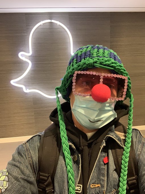 A person wearing a denim jacket, surgical mask, red clown nose, sparkly glasses, and a green braided wig poses in front of a glowing Snapchat neon sign.