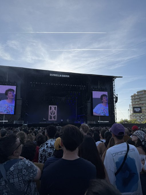 The image shows a large crowd of people gathered at an outdoor event, possibly a concert or festival. They are standing around a stage with two screens displaying images of a man wearing a white shirt and a hat. The audience appears engaged and interested in what is happening on stage. In the background, there are buildings visible, suggesting that this event might be taking place in an urban setting. The post about USDC pairs IRL stands for "International Recording Label", which could potentially be related to the music being played at the event or a sponsorship deal between the event organizers and a USDC label.