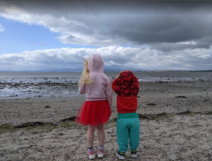 Two children stand with their backs to the camera on a muddy beach looking out at the water under a cloudy sky, one wearing a pink hoodie with a red tutu and the other in a red patterned hoodie with teal pants.