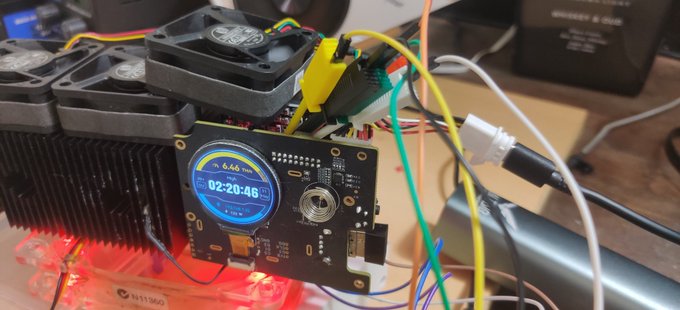 An electronic hardware setup features a small round LCD display showing a timer at '02:20:46' with '6.46' visible, connected to cooling fans, heatsinks, and various colored wires.