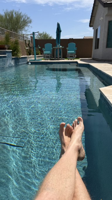 A first-person view shows a person's legs and bare feet with blue-painted toenails dangling over the edge of a swimming pool, with Adirondack chairs and an umbrella visible in the background.