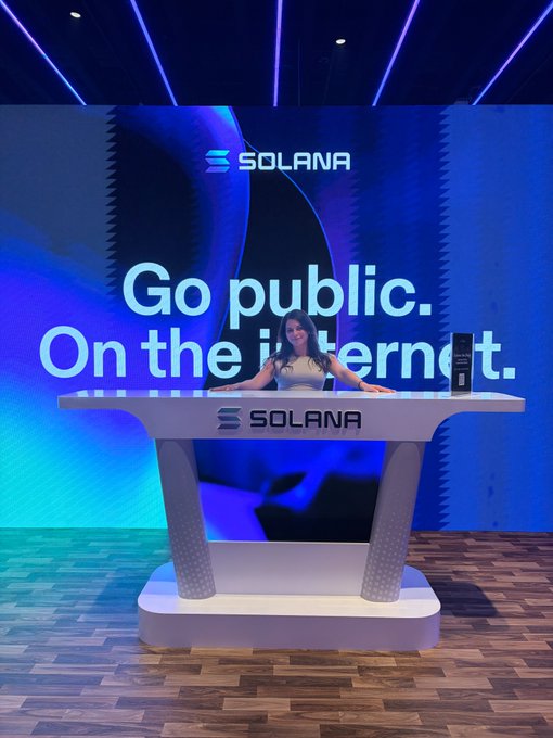 A woman standing behind a Solana-branded podium with a large screen behind her displaying 'Go public. On the internet.'