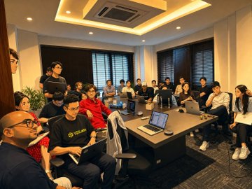 A group of people sitting around a conference table in a meeting room, with laptops open and someone presenting at the front.