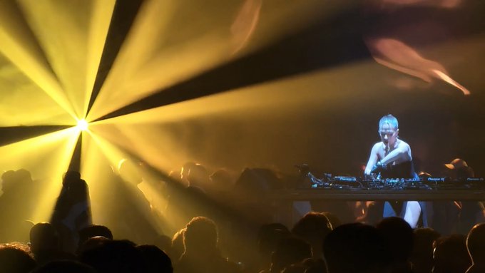 A DJ performs behind decks on stage, illuminated by dramatic yellow spotlights that fan out across the venue while the audience watches in the foreground.