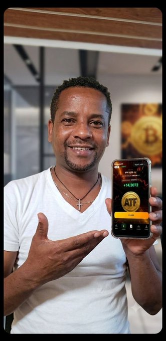 A man wearing a white t-shirt and cross necklace smiles while holding up a smartphone displaying the ATF app, which shows a balance of "+14.9072" in red, set against a background featuring Bitcoin artwork — visually representing his membership in the "$ATF FAMILY."