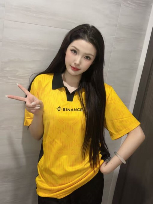 A young woman with long dark hair posing indoors, wearing a yellow Binance-branded jersey and making a peace sign with her hand.
