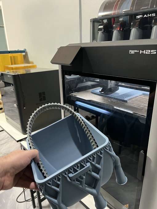 A hand holds a gray 3D-printed basket-like object in front of a Bambu Lab H2S 3D printer in what appears to be a maker space.