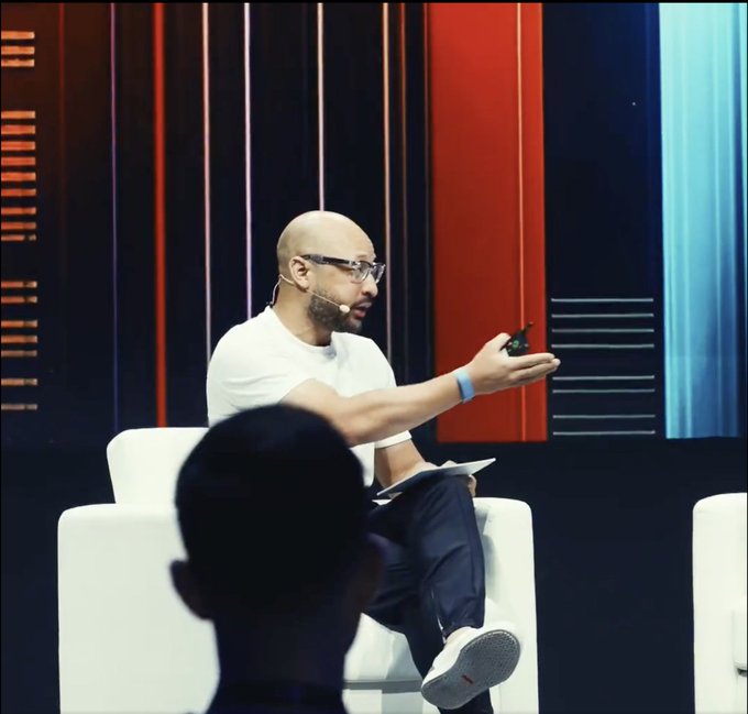 A bald man with glasses sits in a white chair on a stage, speaking into a headset microphone while gesturing with a small device.