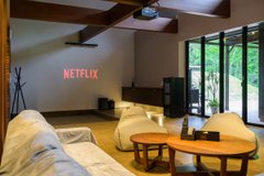A spacious living room features comfortable white sofas arranged around a wooden coffee table, with a projector displaying the "NETFLIX" logo on the wall, suggesting an entertainment setup ideal for a group retreat. The image aligns with the post's description of a newly opened villa in Hidaka, Saitama Prefecture, showcasing its inviting interior as a potential venue for a trading boot camp accommodating multiple guests.