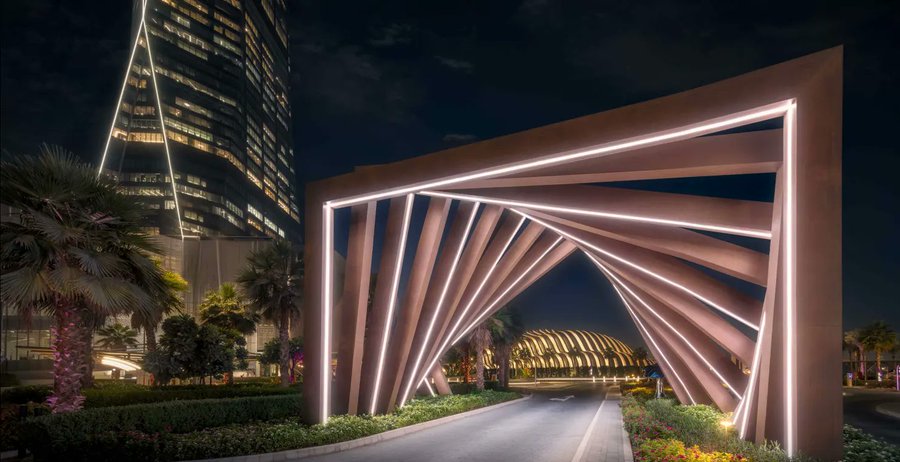 A stunning nighttime view of a futuristic architectural structure with illuminated geometric beams creating a tunnel, set against a backdrop of a tall skyscraper and palm trees.