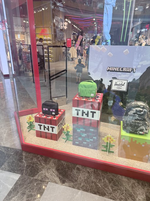 A Minecraft-themed window display features a green Herschel backpack and a black lunch bag on pixelated TNT blocks and grass cubes, with a "MINECRAFT" sign and another Herschel design visible. The setup promotes a collaboration between Herschel and Minecraft, tying into the post's mention of the @vrnouns and @Minecraft edition.