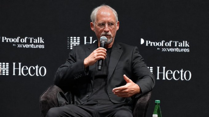 The image features a man sitting down, wearing glasses and a suit jacket. He has his hands together while holding a microphone, suggesting that he might be giving a speech or presentation. The background includes text related to Bitcoin and finance, which indicates that this could be an event or gathering related to the cryptocurrency market or financial discussions.