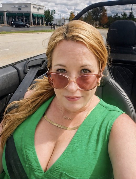 A woman wearing a green top and pink-tinted sunglasses takes a selfie while sitting in a convertible car, with a parking lot and storefronts visible in the background.