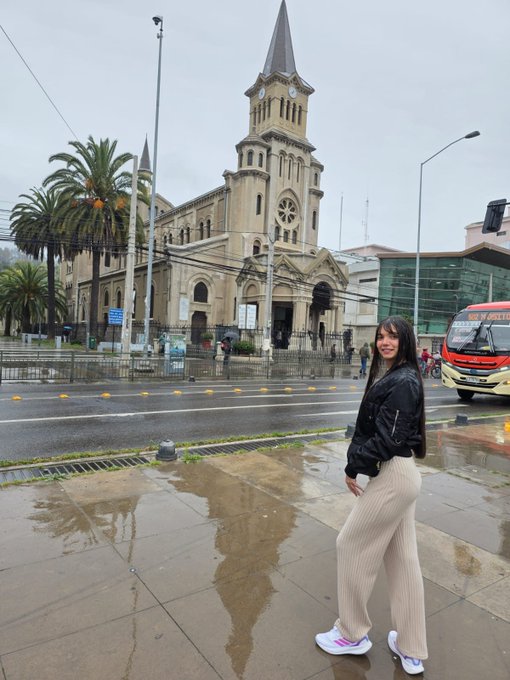 A woman wearing a black jacket, beige pants, and white sneakers stands on a wet sidewalk in front of a large historic cathedral with a tall spire.