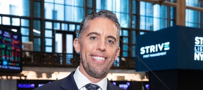 A man in a dark suit and tie smiles for a photo in what appears to be a financial setting, with a sign reading 'STRIVE ASSET MANAGEMENT' visible in the background.