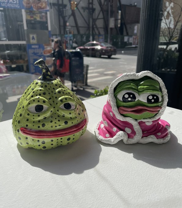 Two Pepe the Frog figurines sit on a white surface near a window; one is shaped like a green pear and the other is wrapped in a pink and white hooded robe.