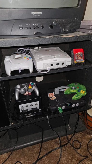 A black entertainment center holding a Curtis Mathes Tronics TV, a Sega Dreamcast console with controllers, and Nintendo 64 consoles with controllers.