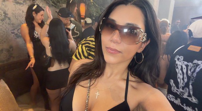 A woman wearing sunglasses and a cross necklace takes a selfie at a crowded indoor event, with another woman in a tiger-striped top visible in the background.