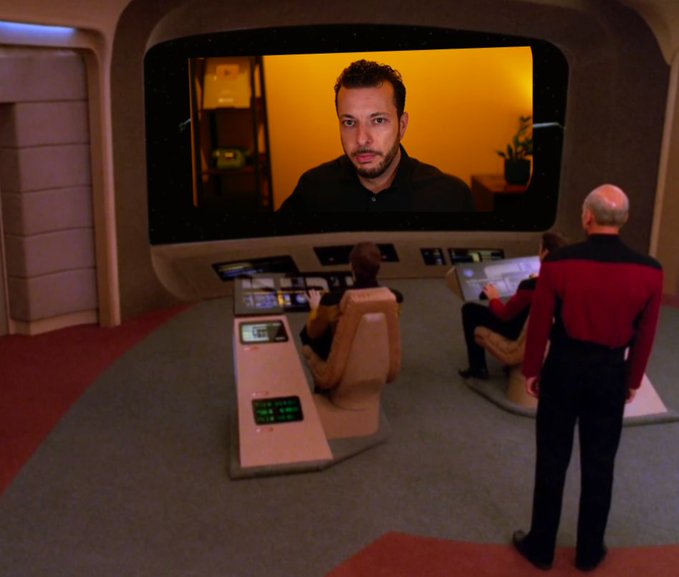 A scene from the bridge of a Star Trek-style spaceship shows crew members in uniform at control consoles, watching a large viewscreen displaying a man with a beard in a dark shirt against a yellow background.