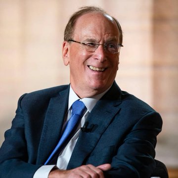 A smiling older man wearing glasses, a dark blue suit, and a blue tie, photographed against a light-colored background.