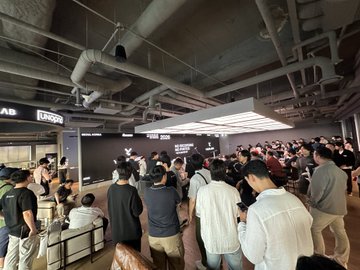 A crowded indoor venue with industrial ceilings and exposed pipes, where people gather around what appears to be a store or kiosk with a 'URBN' sign visible.
