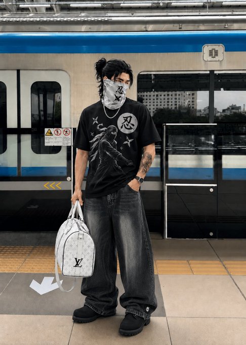 A person with long dark hair wearing a graphic t-shirt, face mask, and dark baggy jeans stands on a subway platform holding a white Louis Vuitton backpack.