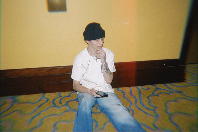 A young man wearing a black beanie and white t-shirt sits on a patterned carpet in a hallway, holding a phone while wearing wired earphones.