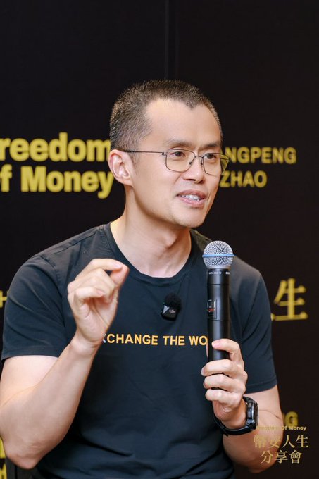 A man with glasses wearing a black 'CHANGE THE WORLD' t-shirt speaks into a microphone at a 'Freedom of Money' event, with 'CHANGPENG ZHAO' visible in the background.