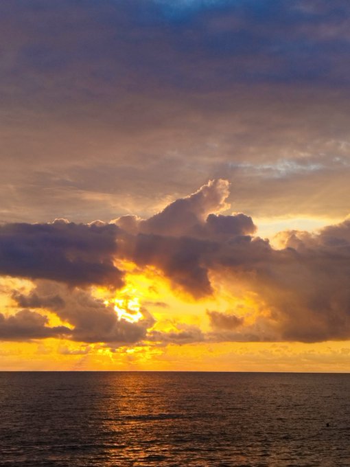 A dramatic sunset over the ocean with golden sunlight breaking through dark, billowing clouds, casting a warm glow across the water.