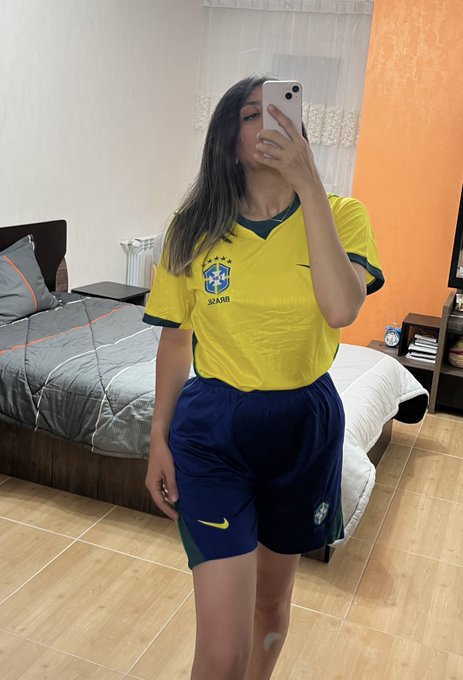 A mirror selfie of a woman wearing a yellow Brazil soccer jersey and navy Nike shorts in a bedroom.