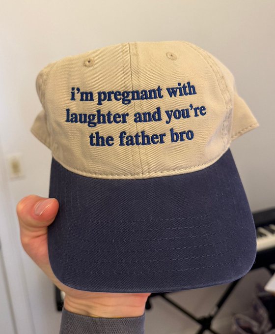 A hand holding a two-tone beige and navy baseball cap with blue embroidered text reading 'i'm pregnant with laughter and you're the father bro'.