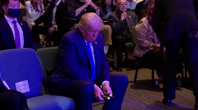 A photograph of Donald Trump sitting in a chair wearing a dark suit and blue tie, holding what appears to be a small object in his hands at what looks like a formal event.