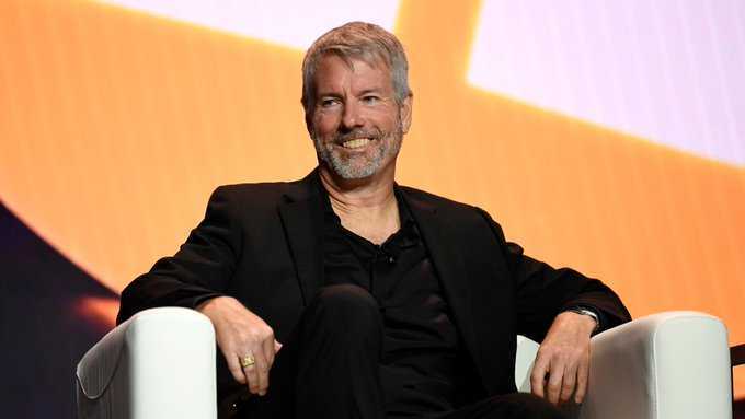 Michael Saylor, MicroStrategy founder, smiling while seated in a white chair against an orange background at an event.