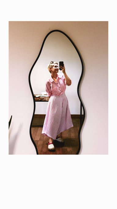 A mirror selfie shows a person wearing a pink and white striped shirt dress, sunglasses, and white shoes, standing in front of a uniquely shaped black-framed mirror.