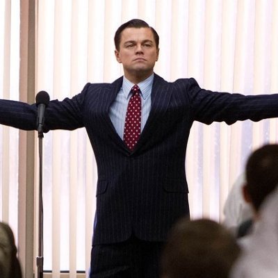 A man in a pinstripe suit and red tie, resembling Leonardo DiCaprio as Jordan Belfort in 'The Wolf of Wall Street', stands with arms outstretched behind a microphone.